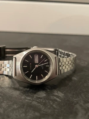 Svart och silver Ricoh herrklocka - Ricoh klocka med svart urtavla och dag/datumvisning. Boett och länk i silverfärgat rostfritt stål. Klassisk design med stilrena indexmarkeringar och länkarmband. Perfekt accessoar för dig som gillar tidlös stil. (Fungerar ej!!, behöver nytt batteri.)