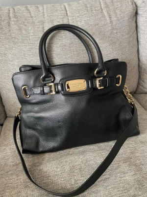 Svart Michael Kors handväska i skinn - Snygg svart handväska från Michael Kors i äkta skinn med guldfärgade metalldetaljer och logoplatta framtill. Väskan har både handtag och axelrem samt flera fack inuti för smart förvaring. Perfekt för dig som vill ha en stilren och lyxig accessoar.