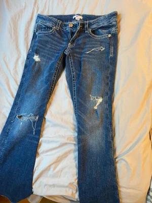 Blå slitna jeans från Gina Tricot - Säljer ett par blå jeans från Gina Tricot med slitna detaljer och hål på benen. Jeansen har klassisk femficksmodell, låg midja och bootcut-ben. Perfekta för en avslappnad och trendig look.