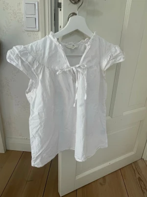 Vit blus med rosett från H&M - Söt vit blus från H&M med korta ärmar och volangdetaljer vid axlarna. Blusen har en liten rosett framtill vid halsringningen och är tillverkad i ett lätt bomullstyg. Perfekt för en fräsch och enkel look.
