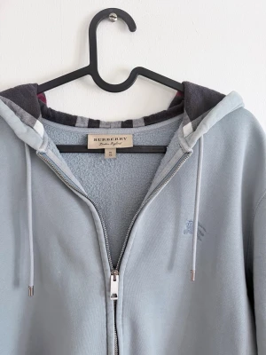 Ljusblå hoodie från Burberry - Snygg ljusblå hoodie från Burberry med dragkedja och klassiskt rutigt foder i huvan. Mjuk insida och diskret logga broderad på bröstet. Perfekt för dig som gillar stilrena och exklusiva plagg med en twist. Ovanlig färg! XL men passar mer som L