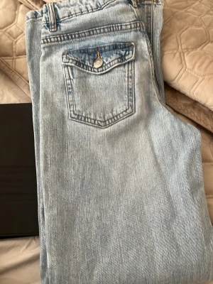 Ljusblå raka jeans med fickdetalj - Säljer ett par ljusblå jeans med rak passform och klassisk femficksdesign. Jeansen har en snygg ficka med knapp bak och är tillverkade i ett mjukt denimtyg. Perfekta för en avslappnad och trendig look. Använt 1 gång 