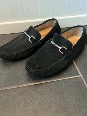 Svarta mockaloafers med metallspänne - Stilrena svarta loafers i mocka med dekorativt metallspänne över vristen. Skorna har klassisk form, låg klack och mjuk innersula för extra komfort. Perfekta för dig som gillar en clean och tidlös look.