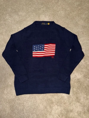 Mörkblå stickad tröja Polo Ralph Lauren - Mörkblå stickad tröja från Polo Ralph Lauren med amerikansk flagga broderad på bröstet och RL-detalj. Tröjan har rund halsringning, ribbade muddar och är gjord i ett mjukt material. Perfekt för en cool och avslappnad stil.