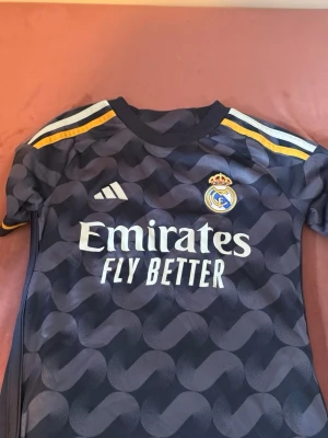 Real Madrid Bellingham matchtröja - Mörkblå Real Madrid matchtröja från Adidas med Bellingham och nummer 5 på ryggen. Tröjan har gula detaljer på axlarna, klubbmärke och sponsortryck framtill samt mönstrad design. Perfekt för fotbollsfans som vill sticka ut på planen eller läktaren.
