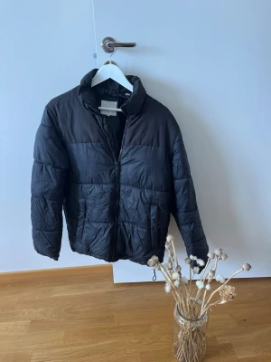 Svart pufferjacka från Jack & Jones - Säljer en svart pufferjacka från Jack & Jones med hög krage och dragkedja framtill. Jackan har två stora fickor på sidorna och quiltad design som ger en schysst streetstil. Materialet är syntetiskt och jackan är perfekt för kalla vinterdagar.