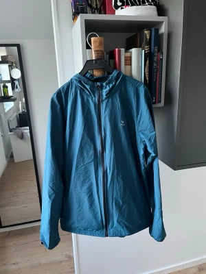 Blå vindjacka från SOC med huva - Snygg blå vindjacka från SOC med hel dragkedja och huva. Jackan har en enkel och stilren design med logga på bröstet. Perfekt för blåsiga dagar och lätt att matcha med jeans eller träningskläder. Väldigt tunn 