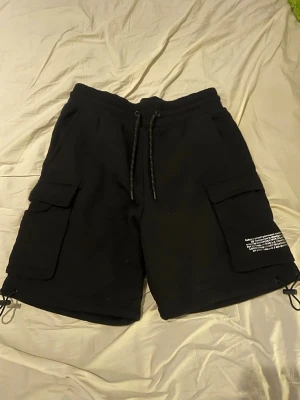 Svarta cargoshorts med snörning - Svarta cargoshorts med stora fickor på sidorna och snörning i midjan samt vid bensluten. Tryckt vit text på ena benfickan ger en cool streetkänsla. Perfekta för en avslappnad och trendig stil.