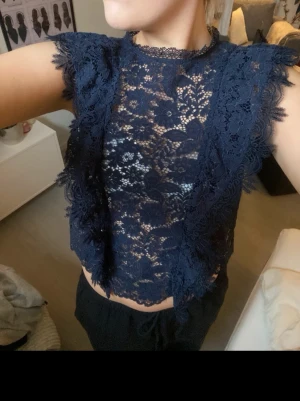 Mörkblå spetsblus med volang - Snygg mörkblå blus i spets med volangdetaljer längs ärmar och framkant. Blusen är ärmlös och har ett blommigt spetsmönster som ger en elegant look. Perfekt att styla med höga jeans eller kjol för en trendig outfit. Det står tyvärr inte vart den är ifrån eller vilken storlek men skulle säga att det är en S💞
