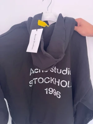  Acne Studios - Snygg mörkgrå hoodie från Acne Studios med tryck 'Acne Studios STOCKHOLM 1996' på framsidan. Klassisk modell med huva och ribbade muddar. Perfekt för en avslappnad och trendig look. Materialet känns mjukt och bekvämt.