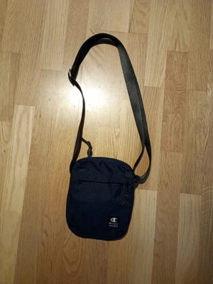 Mörkblå axelväska från Champion - Snygg mörkblå axelväska från Champion med justerbar rem och dragkedja. Väskan har en stilren design med Champion-logga framtill och är tillverkad i slitstarkt tygmaterial. Perfekt för att bära dina viktigaste grejer när du är på språng.