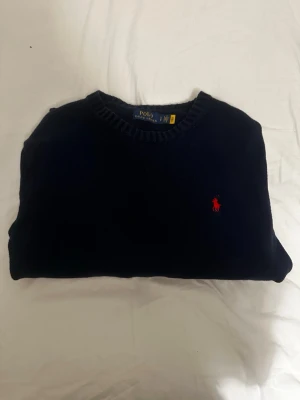 Mörkblå stickad tröja från Polo Ralph Lauren - Mörkblå stickad tröja från Polo Ralph Lauren med klassisk rund halsringning och röd broderad logga på bröstet. Tröjan har en enkel och stilren design som passar perfekt till jeans eller chinos.