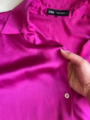 sidenblus från Zara - Snygg fuchsiafärgad blus från Zara i ett glansigt, silkesliknande material. Klassisk krage, knäppning framtill och långa ärmar med manschett. Perfekt för att lyfta vilken outfit som helst med sin starka färg och mjuka känsla.
