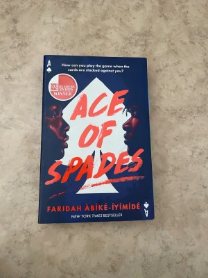 Ace of Spades av Faridah Àbíké-Íyímídé - En ungdomsroman med mörkblått omslag och rödvit text. Boken har en stor vit spader-symbol och ett ansikte i profil på framsidan. Titeln är skriven i stora, röda bokstäver och boken är en New York Times bestseller.