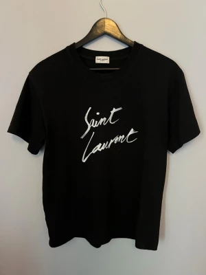 Svart Saint Laurent t-shirt - Svart t-shirt från Saint Laurent med vit logotyp i handskriven stil på bröstet. Klassisk passform med rund hals och korta ärmar. Perfekt för dig som gillar stilrena och trendiga plagg med exklusiv känsla.