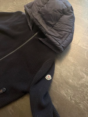 Mörkblå Moncler cardigan med huva - Snygg mörkblå cardiganjacka från Moncler med stickad kropp och quiltad huva. Jackan har två dragkedjeförsedda fickor framtill och Moncler-logga på ärmen. Perfekt för dig som vill ha både stil och komfort i ett plagg. Storlek M, men passar även S! 