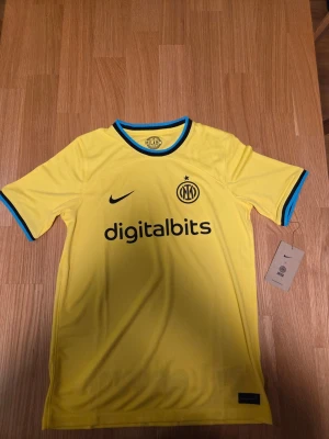 Nike Inter unisex fotbollströja, säsong 22/23 , storlek XL 158-170 cm - Fotbollströja från Inter Milan i gul färg med svarta och blå detaljer. Tröjan har klubbens emblem på bröstet tillsammans med Nike-logotypen och sponsorn Digitalbits. På ryggen finns sponsorn Lenovo tryckt. Tröjan har en rund halsringning och korta ärmar.