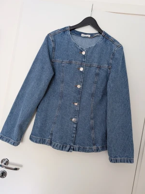 Blå jeansjacka med rund hals från Gina - Snygg blå jeansjacka med figursydd passform och rundad halsringning. Jackan har långa ärmar och fem silverfärgade knappar framtill. Klassisk denimlook utan krage och utan synliga fickor, perfekt för en clean och stilren outfit. Storlek L. Aldrig använd. 
