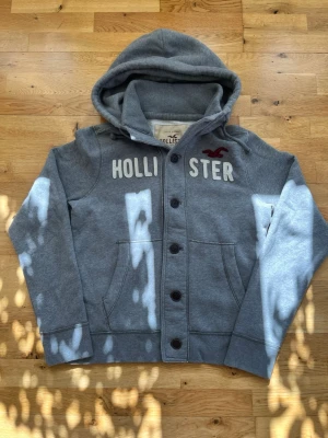  Vintage Hollister Cardigan hoodie med knappar - Säljer nu denna snygga grå vintage  Hollister cardigan. Tröjan har vit Hollister text broderat på framsidan, samt en vinröd Hollister logga. Står att tröjan är i storlek XL, men passar som L/M.  Skick 9/10.  Längd: 68 cm Bredd (armhåla till armhåla): 55 cm Skriv gärna om ni har några mer funderingar🙂