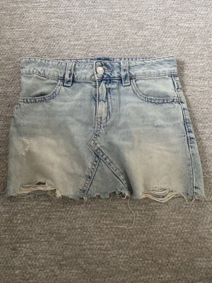 Ljusblå jeanskjol - Cool ljusblå jeanskjol med rå, fransig kant och slitna detaljer. Kjolen har klassiska fickor fram och bak, samt snygga broderier på bakfickorna. Perfekt för dig som gillar en avslappnad och trendig stil.
