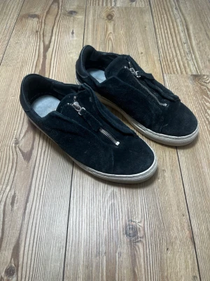 Axel Arigato skor med dragkedja - Svarta sneakers i mocka med cool dragkedja framtill istället för klassiska snören. Skorna har vit sula och en stilren, minimalistisk design som passar till det mesta. Perfekt för dig som gillar en clean look med lite edge. I använt skick och saknas sula! I mocka