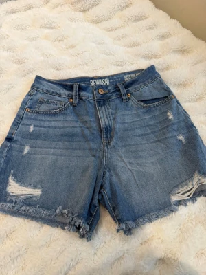 Blåa high waist jeansshorts Rewash - Säljer ett par blåa högmidjade jeansshorts från Rewash med slitna detaljer och fransig kant. Klassisk femficksmodell med knapp och dragkedja. Perfekta till sommaren för en avslappnad och trendig look.