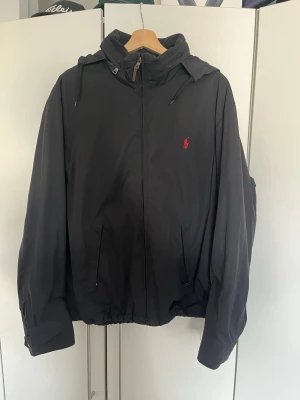 Svart vindjacka från Polo Ralph Lauren - Svart vindjacka från Polo Ralph Lauren med huva och dragkedja framtill. Jackan har två fickor med knappar och en röd broderad logga på bröstet. Perfekt för blåsiga dagar och enkel att matcha med olika outfits.