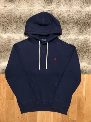 Marinblå Polo Ralph Lauren Hoodie - Storlek S - Hej! 👋 Säljer en riktigt snygg marinblå Polo Ralph Lauren hoodie i storlek S med röd pony-logga. 🐎 Tröjan är i mycket fint skick. Det finns en liten mörkare prick på magfickan (se sista bild), men det är inget som stör helhetsintrycket när den bärs. ✨ 100% äkta med originaltvättlappar kvar och en fullt fungerande QR-kod för enkel verifiering ✅ Ett perfekt premium-basplagg till garderoben! Fraktar snabbt! 🚛📦💬 