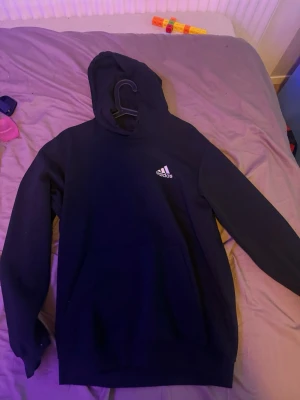Svart hoodie från Adidas - Svart hoodie från Adidas med klassisk vit logga på bröstet. Hoodien har huva och en stor magficka framtill. Perfekt för en avslappnad och sportig stil. Mjuk och skön bomullsblandning som passar till det mesta.