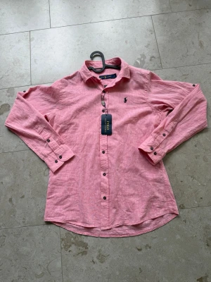 Rosa linneskjorta från Polo Ralph Lauren - Snygg rosa skjorta från Polo Ralph Lauren i linne med klassisk krage och knappar framtill. Skjortan har lång ärm och ikonisk broderad logga på bröstet. Perfekt för en fräsch och avslappnad stil.