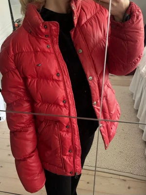 Röd dunjacka från Tommy Hilfiger - Snygg röd pufferjacka från Tommy Hilfiger med hög krage, tryckknappar och dragkedja. Jackan har två stora fickor framtill och diskret logga på bröstet. Perfekt oversized look och riktigt varm för kalla dagar. Materialet är glansigt och quiltat för extra stilpoäng. Storlek L men passar mig som är xs/s