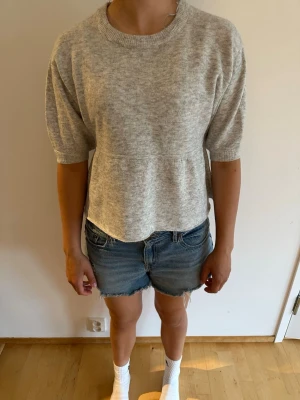 Ljusgrå stickad topp med volang Nelly - Säljer en gullig ljusgrå stickad topp med rund halsringning och korta ärmar. Toppen från Nelly har en volang och är tillverkad i mjukt, tunt stickat material. Perfekt att styla med jeans eller kjol för en chill look.
