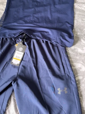 Blått träningsset från Under Armour - Säljer ett blått träningsset från Under Armour med matchande tröja och byxor. Setet har en sportig look med elastisk midja på byxorna och fickor. Tillverkat i ett lätt och funktionellt material, perfekt för träning. Under Armour-logga på byxbenet.