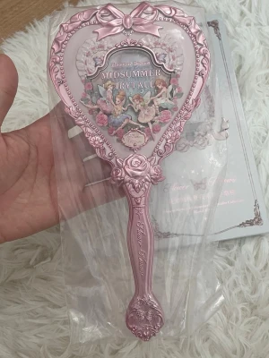 Midsummer Fairytale handhållen spegel - Supersöt spegel från Flower Knows i hjärtformad design. Spegeln är ljusrosa med detaljer som rosetter och rosor samt ett sagolikt motiv med älvor och blommor. 