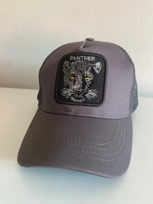 Grå keps med panterpatch - Snygg grå keps med svart mesh baktill och en broderad patch framtill med motiv av en panter och texten 'PANTHER'. Kepsen har böjd skärm och justerbar snapback. Perfekt för dig som gillar djurmotiv och streetwear.