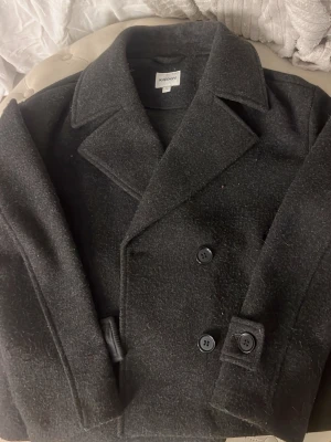 Svart/grå  kappa - Kappan har långa ärmar och stora knappar framtill, perfekt för kyliga dagar. Enkel och tidlös design som passar till många outfits.