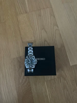Seiko  - Stilren Seiko gmt master klocka med silverfärgat metallarmband och svart urtavla. Klockan har tydliga siffror och markeringar runt boetten samt en robust design. Perfekt accessoar för dig som gillar klassisk och tidlös stil.