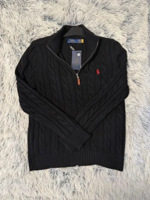 Svart tröja från Polo Ralph Lauren - Kabelstickad svart tröja från Polo Ralph Lauren med halv dragkedja och ribbade muddar. Klassisk röd logga broderad på bröstet och hög krage. Perfekt för dig som gillar stilrena och tidlösa plagg med premiumkänsla.
