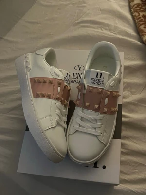 Valentino Rockstud Untitled sneakers - Snygga vita sneakers från Valentino med breda ljusrosa band över vristen dekorerade med ikoniska pyramidnitar i guld. Skorna har en chunky vit sula och klassisk låg siluett. Perfekta för dig som vill ha en unik och trendig look.