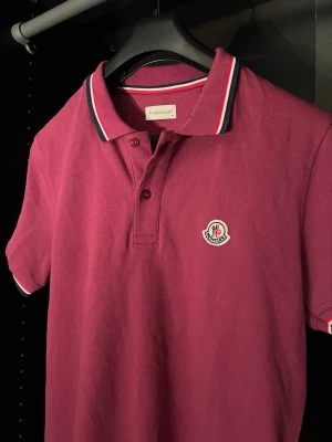 Lila pikétröja från Moncler - Snygg Lila pikétröja från Moncler med klassisk krage och knappar framtill. Tröjan har vita och marinblå detaljer på krage och ärmslut samt Moncler-logga på bröstet. Tillverkad i mjuk bomull och har korta ärmar