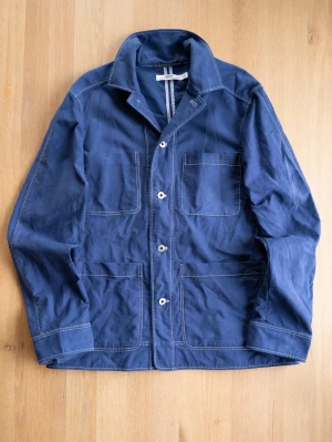 Marinblå Chore Jacket från Hope, size M - Snygg marinblå overshirt från Hope i robust bomullstyg. Jackan har vita kontrastsömmar, tre stora utanpåliggande fickor och knäppning framtill. Perfekt lager-på-lager-plagg med avslappnad passform och klassisk krage.