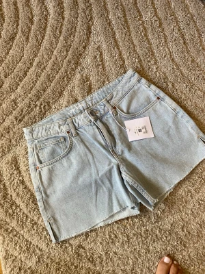 Ljusblå denimshorts från Bik bok - Säljer ett par ljusblå denimshorts från Monki med klassisk femficksdesign och rå kant nedtill. Shortsen har hög midja och stängs med dragkedja och knapp. Perfekta för varma dagar och enkel att matcha med allt.