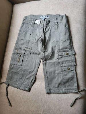 Khaki cargobyxor med fickor Wet Seal storlek 0 USA XS - Säljer ett par khaki cargobyxor med flera stora fickor, dragkedjedetaljer och knappar. Byxorna har snörning vid bensluten och breda hällor för bälte. Tillverkade i bomull för en skön och avslappnad look.
