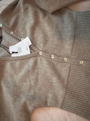 Beige ribbad kofta från Gina Tricot - Säljer en beige, ribbad kofta från Gina Tricot med v-ringning och knappar framtill. Koftan har en figurnära passform och bred ribbad kant nedtill. Perfekt att styla över ett linne eller t-shirt för en chill look.