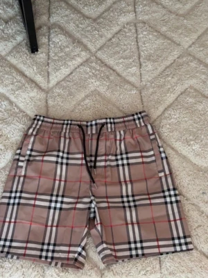 Beige rutiga shorts med snörning - Säljer ett par beige shorts med klassiskt rutigt mönster och svarta snören i midjan. Shortsen har elastisk midja och fickor på sidorna samt bak. Perfekta för sommaren och riktigt snygga till en enkel t-shirt. Dom är i L men skulle säga att dom passar dig som har M bättre 