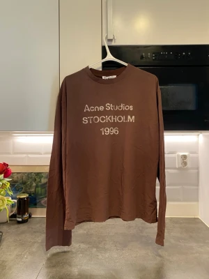 Brun långärmad topp Acne Studios - Cool brun långärmad topp från Acne Studios med tryck 'Acne Studios STOCKHOLM 1996' på bröstet. Tillverkad i mjuk bomull med relaxed fit och rund hals. Perfekt för dig som gillar minimalistisk och stilren design.
