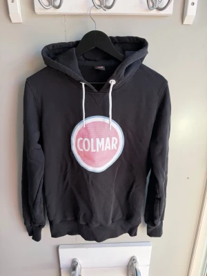 Svart Colmar hoodie med tryck - Snygg svart hoodie från Colmar med stort rosa och vitt logotryck på bröstet. Klassisk huva med vita dragsnören och ribbade muddar vid ärmar och nederkant. Perfekt för en avslappnad och trendig stil. Mjuk bomullskänsla.