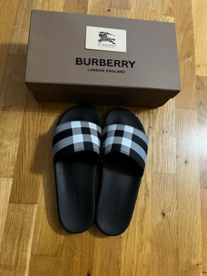 Burberry Furleys Svart - Säljer ett par snygga slides från Burberry med klassiskt svartvitt rutmönster över foten. Sandalerna har en svart sula i gummi och bred rem med Burberrys ikoniska design. Perfekta för en trendig och avslappnad stil. Storlek 44