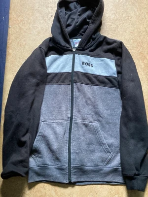 Grå och svart hoodie från BOSS - Säljer en grå och svart hoodie från BOSS med dragkedja och huva. Hoodien har broderad BOSS-logga på bröstet, två stora fickor framtill och är tillverkad i mjukt material som känns skönt mot huden. Perfekt för dig som gillar en avslappnad och cool stil.