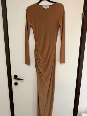 Brun långklänning med rynkdetalj - Säljer en brun långklänning med lång ärm och rund halsringning. Klänningen har en snygg rynkdetalj på sidan som ger en cool draperad effekt.  Den har en slits på sidan av benet Tillverkad i ett mjukt och stretchigt material som sitter snyggt på kroppen.